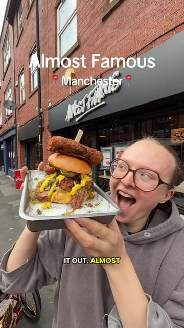 A high-energy, vlog-style review of a 'pickle burger' and various sides at Almost Famous in Manchester. The creator showcases the indulgent, saucy nature of the food, including mac and cheese balls, tater tots, and a cocktail, while maintaining an enthusiastic tone throughout.