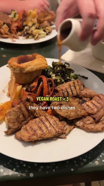 A creator reviews a vegan Sunday roast at a bakery/cafe, highlighting the 'beef' wellington and 'chicken' pieces, along with classic trimmings like Yorkshire puddings and cauliflower cheese. The video includes shots of the food, the venue's interior, and mentions the dog-friendly policy.