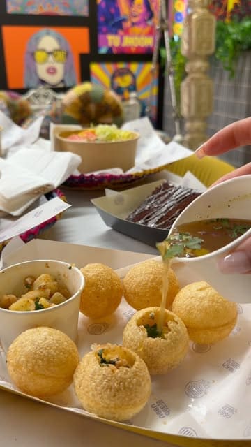 A montage of various dishes including loaded fries, golgappe, and naan, set within a vibrant, colorful restaurant space featuring a tuk-tuk.
