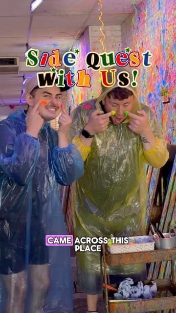A high-energy, vlog-style video featuring two creators visiting a 'Paint Splat' venue in Manchester. They demonstrate various methods of painting, including using fly swatters, syringes, water guns, and a spin art turntable, while wearing protective ponchos.