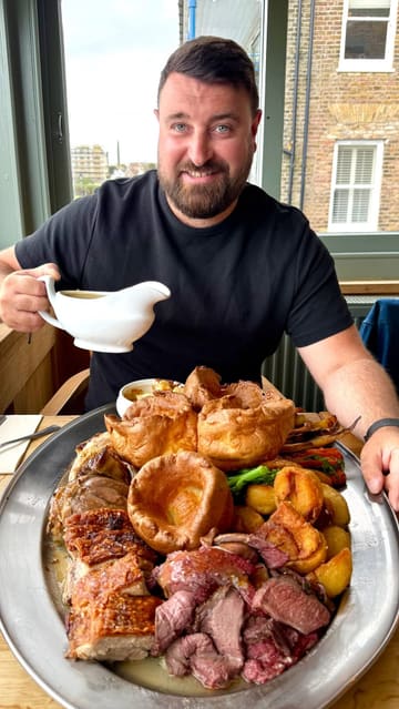 A short, visually appealing clip featuring a man pouring gravy over a large sharing platter of Sunday roast meats, including beef, chicken, lamb shank, and pork belly, accompanied by Yorkshire puddings and roast potatoes.