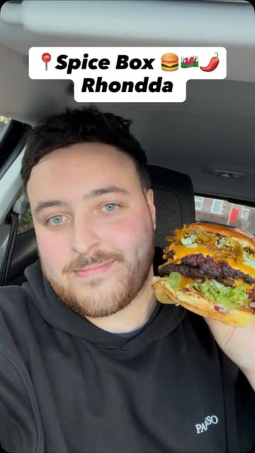 A creator reviews a large 'munch box' from Spice Shack, a takeaway operating out of a rugby club in Rhondda. The video features close-ups of various Indian-fusion items including burgers, chicken tenders, tandoori wings, and sides, with the creator providing commentary on the flavours.
