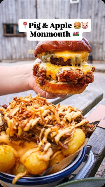 A high-quality, appetizing close-up shot of a thick, juicy burger held in hand, positioned above a bowl of loaded tots. The rustic, outdoor farm setting provides an authentic backdrop that complements the casual, indulgent nature of the food.