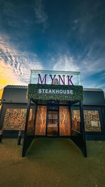 A promotional montage for Mynk Steakhouse featuring shots of the restaurant's interior decor, neon signage, mocktails, and various meat dishes including lamb chops, prawns, and steaks.