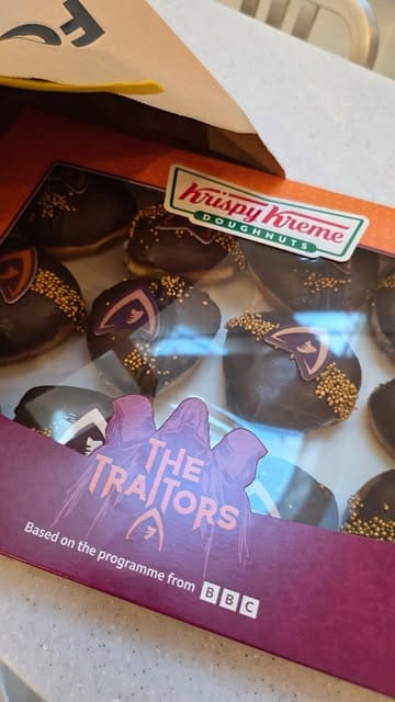 A short, handheld video showcasing a box of 'The Traitors' themed Krispy Kreme doughnuts, featuring a close-up reveal of the different coloured fillings.