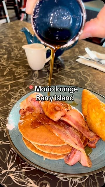 A short montage video showcasing the Morio Lounge in Barry Island, featuring close-ups of a full breakfast and buttermilk pancakes with maple syrup, alongside shots of the venue's interior decor and the creator enjoying an iced coffee.