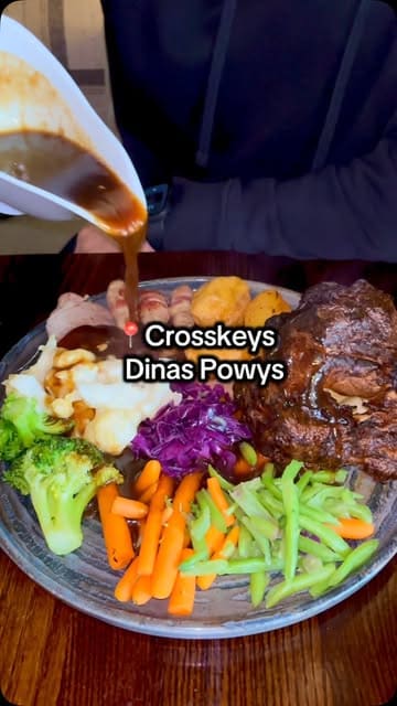 A couple visits The Cross Keys in Dinas Powys to review their three-course Sunday roast, showcasing the starters, main course, and desserts while highlighting the affordable price point.
