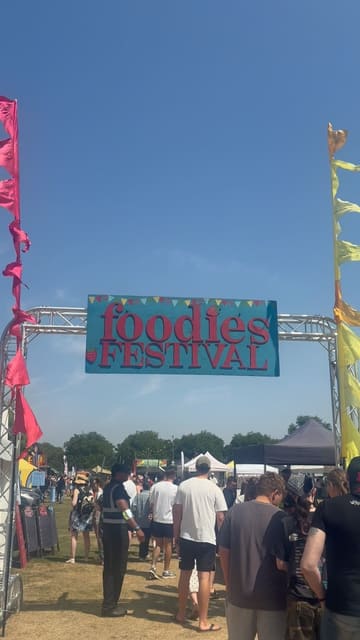 A montage of various food and drink items available at the Foodies Festival, including gyros, a burger, dumplings, ice cream, and various drinks, interspersed with shots of the festival grounds.