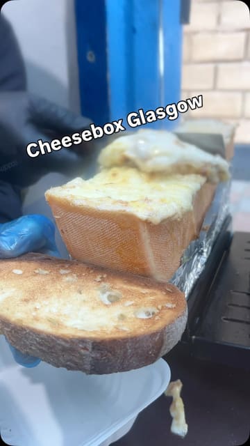 A short-form video review of Cheese Box Glasgow, a street food stall located in a converted police box. The creator walks through the menu, showing the preparation of raclette, cheesy bagels, cheesecake, and hot chocolate, providing a clear and appetizing overview of the offerings.