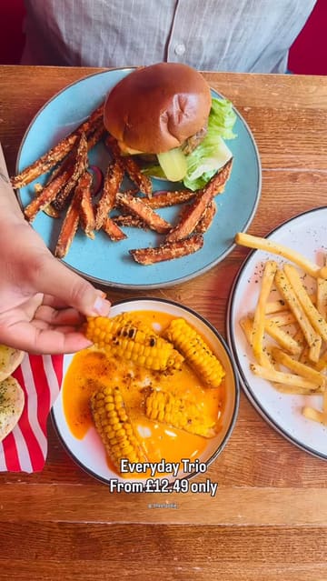 A short, energetic video showcasing a visit to TGI Fridays, highlighting their 'Everyday Trio' menu deal. The creator captures the exterior, the menu, the interior decor, and various food and drink items, including burgers, fries, corn, and cocktails.