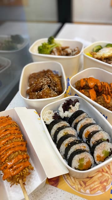A casual, handheld video showcasing a visit to Bunsik, featuring shots of the exterior, the ordering area, and a variety of Korean dishes including corn dogs, fried chicken, rice boxes, and kimbap.