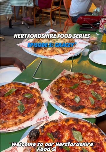 Two creators visit Gracey's Pizza in St Albans as part of their Hertfordshire food series. They show the exterior, the kitchen where dough is prepared, the menu, and enjoy a meal of olives, various pizzas, and cheesy garlic bread at an outdoor table.