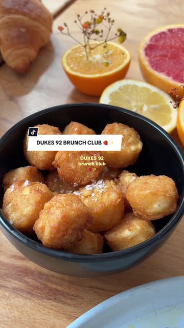A creator visits Dukes 92 in Manchester to review their new brunch club offering. The video features shots of the venue's interior and outdoor seating, the brunch menu, coffee, cocktails, and food items like poached eggs on focaccia and a fruit and pastry platter.