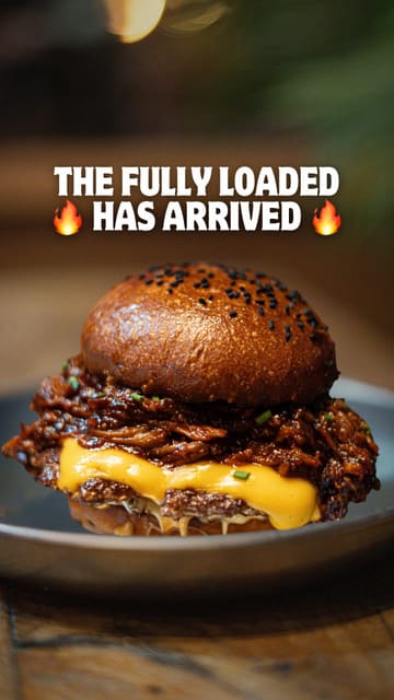 A high-quality, close-up food photography shot of a 'fully loaded' burger featuring double smashed patties, cheese, and pulled pork, presented on a dark plate with text overlay.