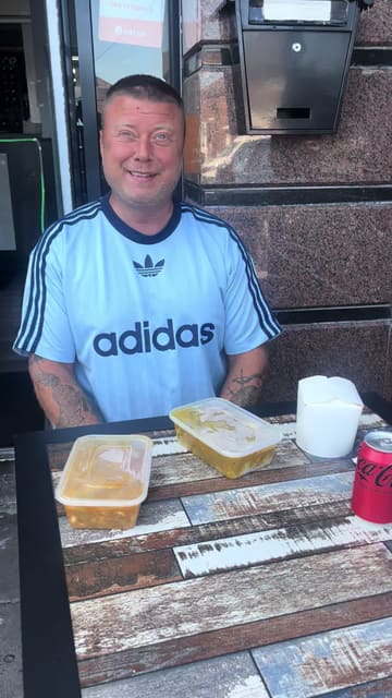 A man reviews Chinese takeaway food, specifically a crispy duck noodle box, chicken satay, and spring rolls, while sitting at an outdoor table.