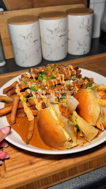 A short, top-down recipe video demonstrating how to make a 'dipped burger' meal at home using Nando's branded sauce and peri-salt. The video shows the assembly process, including dipping the burger, adding fries, and garnishing with various toppings.