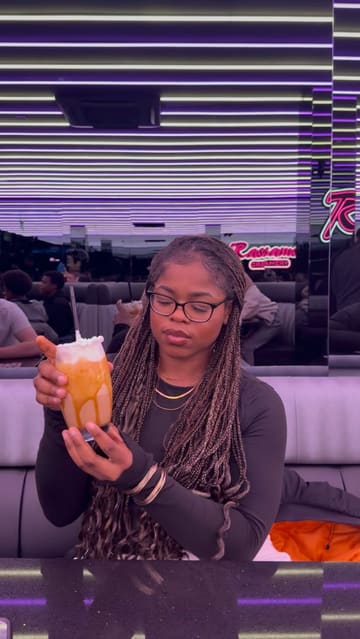 A creator visits Rassams Creamery in Manchester, showcasing the extensive dessert menu, the robot server, and the vibrant, neon-lit interior. The video features a review of their waffles and crepes, highlighting the indulgent nature of the food.