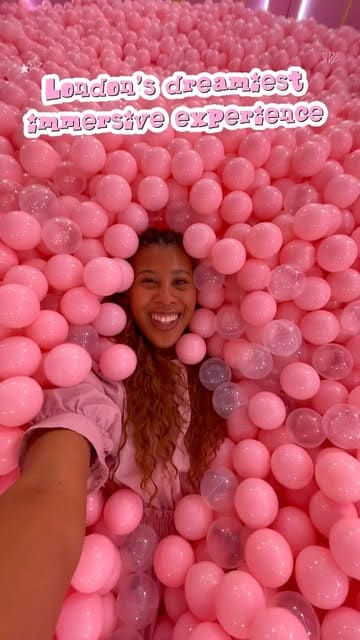 A bright, selfie-style shot of a creator immersed in a pink ball pit at the House of Dreamers exhibition in London. The image effectively communicates the fun, interactive nature of the venue.