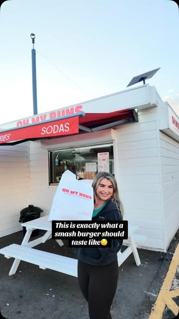 A creator visits a burger takeaway spot called Oh My Buns, showcasing the menu, the smash burgers, fries, milkshakes, and cookies, and provides a positive review while eating in her car.
