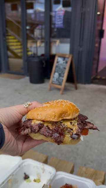 A casual, first-person perspective shot of a smash burger being held up, likely at a street food venue. The focus is on the burger, showcasing its messy, juicy texture, with a blurred background suggesting an outdoor dining setting.