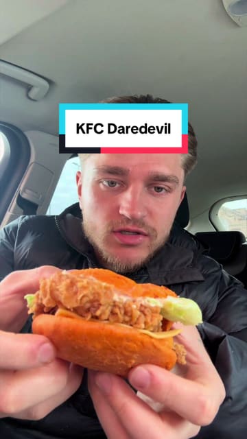 A creator reviews the limited-edition 'Daredevil' menu from KFC, specifically testing the loaded fries, chicken burger, and hot wings while sitting in a car. The video features direct-to-camera commentary and close-up shots of the food items.