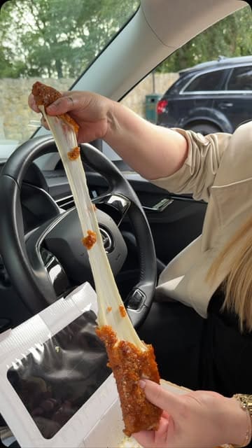 A food review video featuring a creator trying various indulgent takeaway items like mozzarella sticks, tacos, chicken tenders, and loaded fries from a venue called 'The Hunger' while sitting in a car.