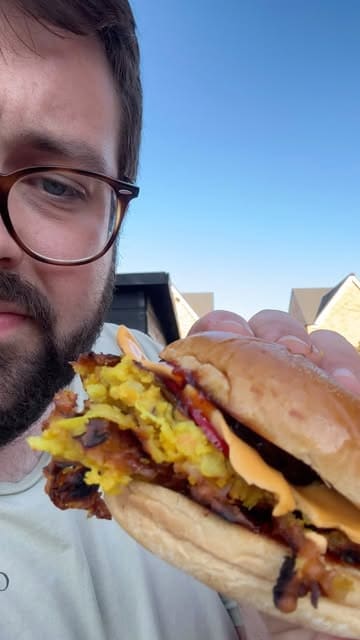 A home cooking video where the creator demonstrates how to make an onion bhaji smash burger on a BBQ grill, inspired by a poor experience with a local Indian takeaway.
