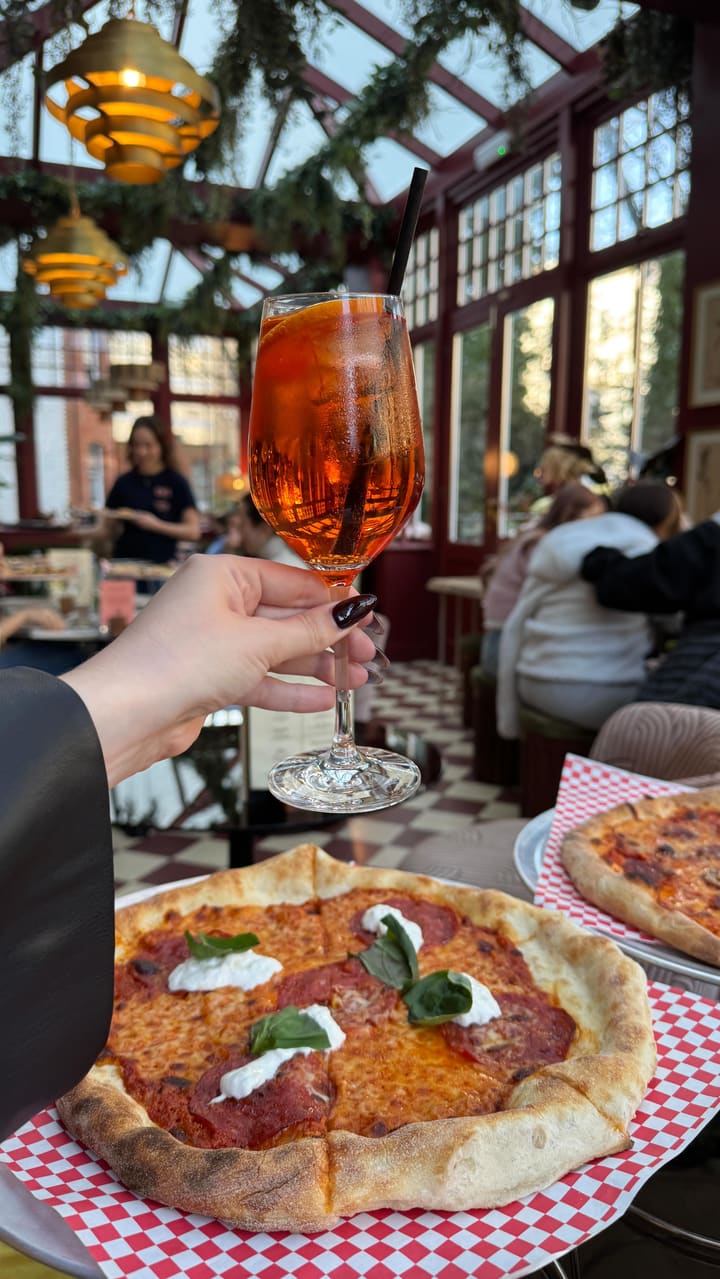 The video showcases the EVE X Proper Slice restaurant, highlighting its interior, food, and drinks. The setting is a stylish restaurant with a conservatory-like area, featuring hanging plants and unique lighting fixtures. The food shown includes pepperoni pizza topped with dollops of white cheese and basil, a Caesar salad with anchovies and chicken, and a cocktail. The pizza is presented on a red and white checkered paper. The video focuses on the dining experience and the restaurant's ambiance.