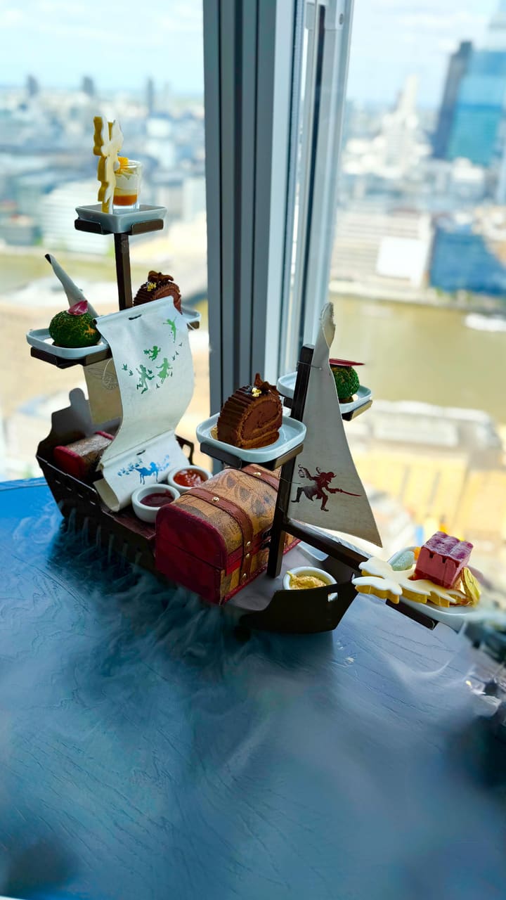 The video showcases a Peter Pan-themed afternoon tea experience at Aqua Shard in The Shard, London. The tea is presented on a multi-tiered ship-shaped stand with dry ice creating a smoky effect. It features various sweet and savory items, including sandwiches served on a clock-face plate, scones presented in a treasure chest, and themed desserts. The video also highlights the panoramic views of London from the venue and shows glasses of champagne and cups of tea. It mentions that a donation is made to Great Ormond Street Hospital for every afternoon tea purchased.