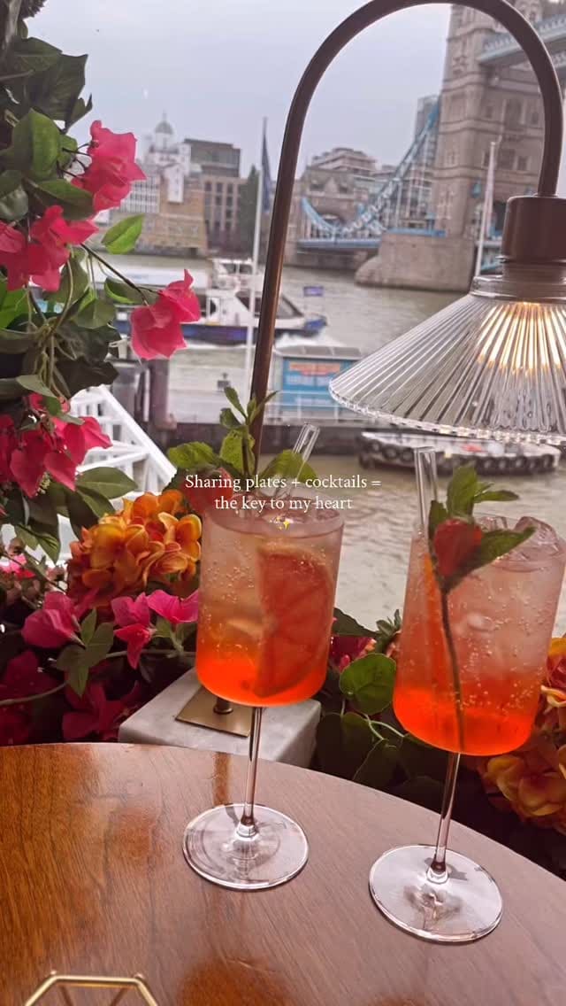 A montage-style video showcasing a dining experience at a restaurant with a prominent view of Tower Bridge in London. The video features several small plates (tapas-style), including fried chicken bites, garlic prawns, meatballs in a white sauce, fried cheese or risotto balls, and a caprese-style salad with tomato, mozzarella, and avocado. It also shows two red/orange spritz-style cocktails. The video intersperses shots of the food with scenic views of the River Thames and Tower Bridge, as well as a brief shot of the restaurant's bar area.