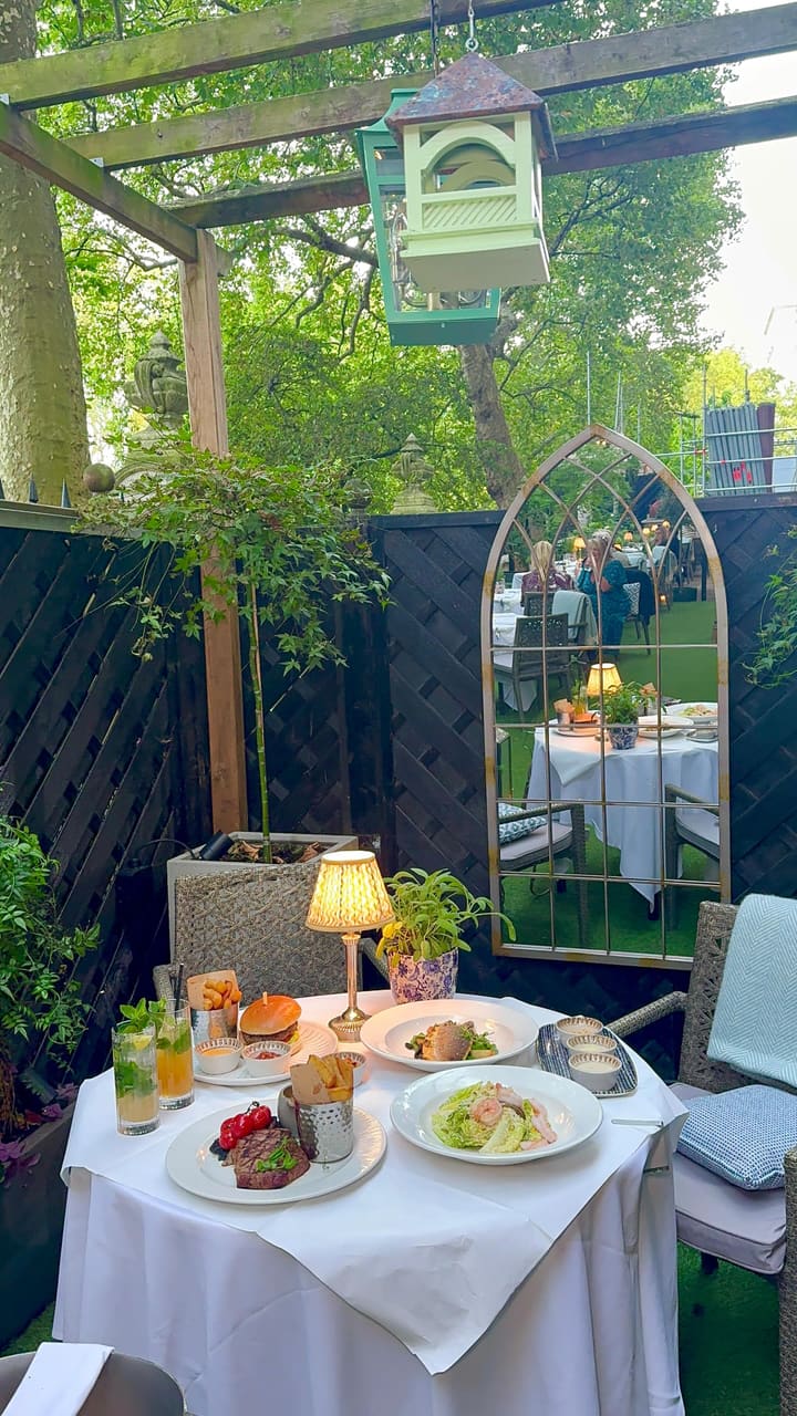 The video promotes the outdoor terrace restaurant at The Royal Horseguards Hotel in London. It showcases the elegant and green-filled dining space during the day and evening. Various dishes are featured, including a steak with fries and tomatoes, a pan-seared sea bass, a shrimp Caesar salad, and a burger. There are dynamic action shots of Caesar dressing being poured on the salad and a creamy sauce over the steak. A dessert with torched meringue is also shown. The video concludes with shots of the hotel's luxurious interior and grand exterior.