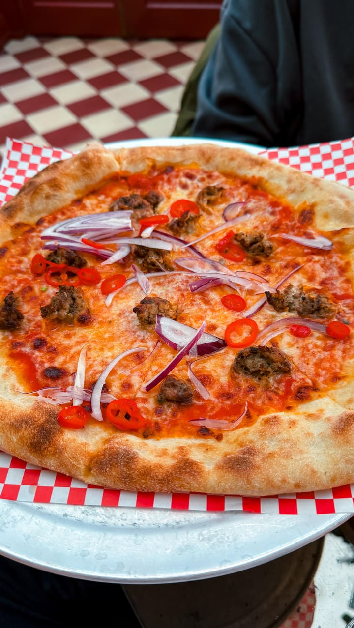 The video showcases Eve Kensington, a restaurant in Earl's Court, London, specializing in NYC-style pizza. The setting includes indoor and outdoor seating areas with stylish decor. The food and drinks featured are Pepperoni Pizza, Mafioso Pizza (with sausage, red onion, and chili), Mac & Cheese, an Aperol Spritz, and beer. The pizzas are presented on red and white checkered paper on metal stands. The Mac & Cheese is served in a small white bowl. The video focuses on highlighting the restaurant's ambiance and the food's visual appeal.