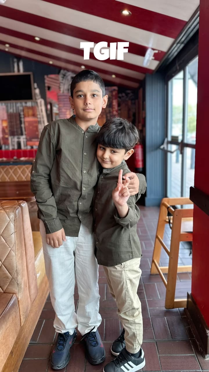 The video showcases a family's visit to a TGI Fridays restaurant. The main focus is the 'Dubai Chocolate Milkshake,' a light green milkshake served in a tall glass, topped with whipped cream, chocolate sauce, and a rim coated in chocolate and shredded phyllo pastry (kataifi). The video features a woman and two young boys enjoying the milkshakes and other food items, including spicy chicken wings, a burger with fries, and nachos. The setting is a classic American-style diner with red leather booths and themed decor.