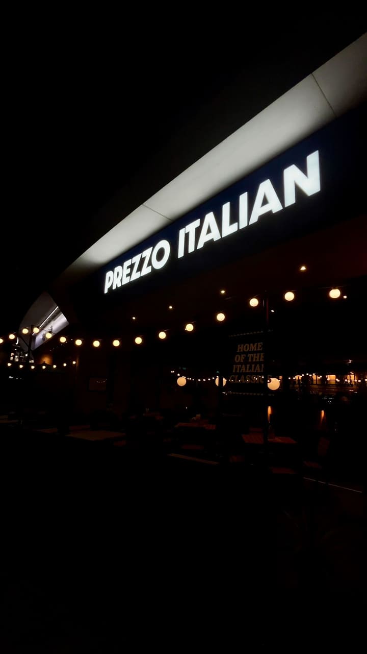 The video showcases a dining experience at Prezzo, an Italian restaurant in Chelmsford. It begins with exterior shots of the restaurant at night, highlighting its signage. The video then moves inside, showing the cozy, modern interior, a cocktail, and a gluten-free menu. Several dishes are featured in close-up shots, including a burrata and tomato salad with a 'Gluten-Free' flag, a bowl of olives, a creamy chicken and mushroom fusilli pasta, a baked lobster mac and cheese, and truffle fries. The video concludes by showing the dessert menu and a slice of chocolate fudge cake with vanilla ice cream, which has a celebratory sparkler in it.