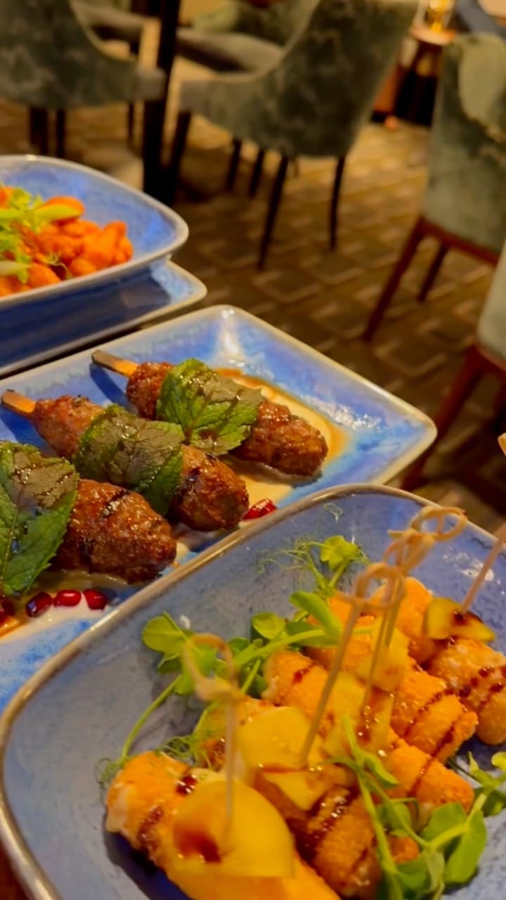 The video promotes Archy's Bar & Lounge. It begins with a close-up of the menu, then showcases a variety of small plates served on blue dishes. These include what appear to be fried chicken or fish bites with a dipping sauce, lamb kofta skewers garnished with mint, and breaded cheese sticks with apple slices. A person is shown breaking one of the cheese sticks to reveal a cheese pull. The video also features a main course of spaghetti with chicken and vegetables. The final shots show the interior of the lounge, which includes a large TV screen over a fireplace showing a sports game.