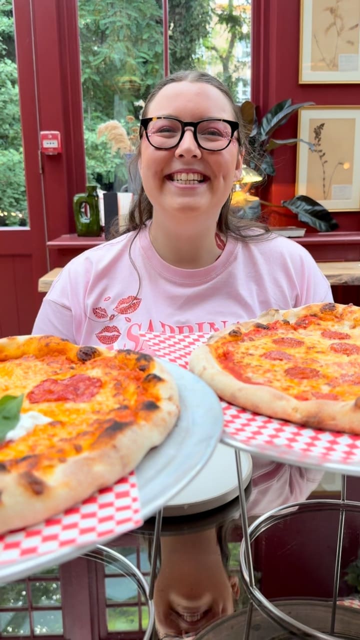 The video showcases a visit to a stylish restaurant, Eve Kensington. It features a woman happily receiving two large pizzas. There are multiple shots of the restaurant's interior, which has a conservatory-like feel with large windows, red-painted frames, checkered floors, lush plants, and eclectic, colourful furniture. The food shown includes two types of pizza (one pepperoni, one with salami and burrata), and a side of parmesan fries. Key food moments include a cheese pull, dipping the pizza crust into a sauce, and taking a bite. The overall vibe is fun, positive, and aesthetically pleasing.