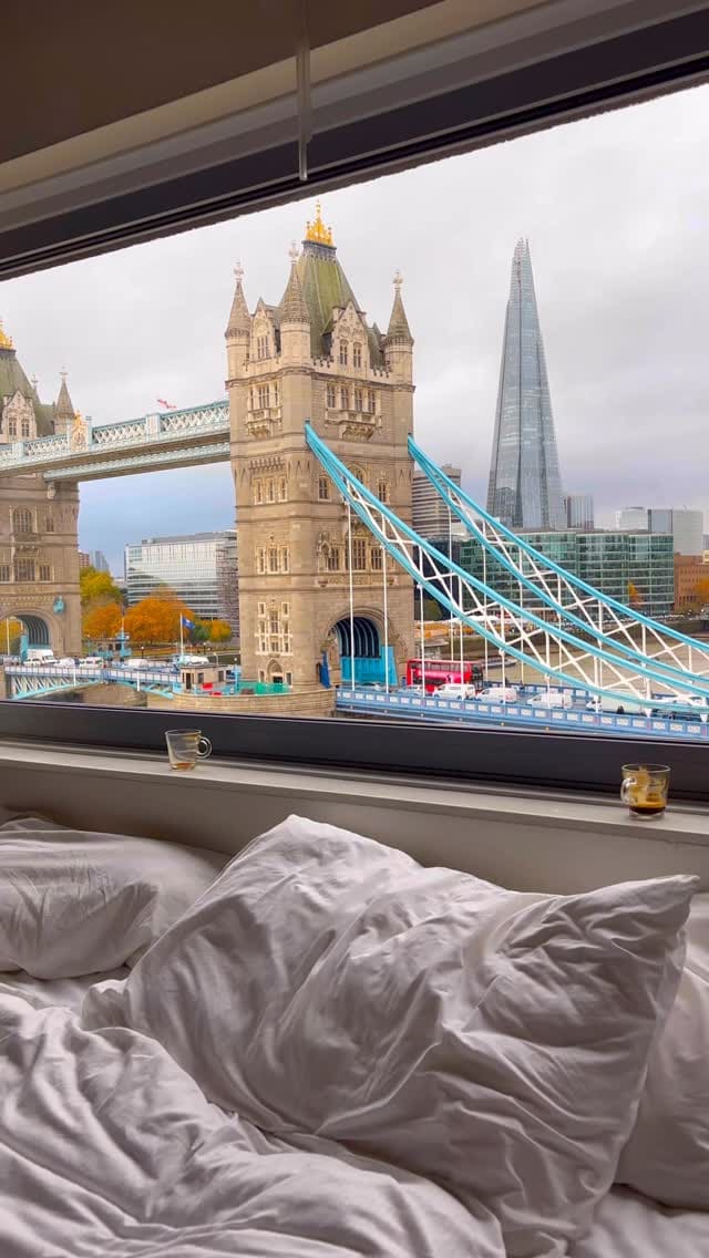The video showcases a hotel room with a direct, stunning view of the Tower Bridge and The Shard in London. It features various shots of the view from the window during the day and at night. A woman is shown relaxing in a bathrobe, looking out the window, and sleeping in the comfortable bed, conveying a luxurious and relaxing stay.