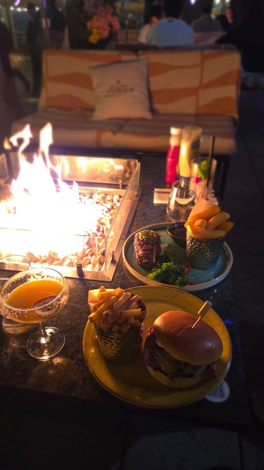 The video showcases a nighttime dining experience at Vicinity, a restaurant in London. It highlights the outdoor seating area with fire pits and a clear view of the illuminated Tower Bridge. The video features various food and drink items, including two cocktails (a passionfruit martini and a red berry cocktail), small plates like gochujang popcorn chicken and another fried dish, a 10oz sirloin steak with fries and kale, and a large burger with fries. The overall atmosphere is presented as 'laidback luxury.'