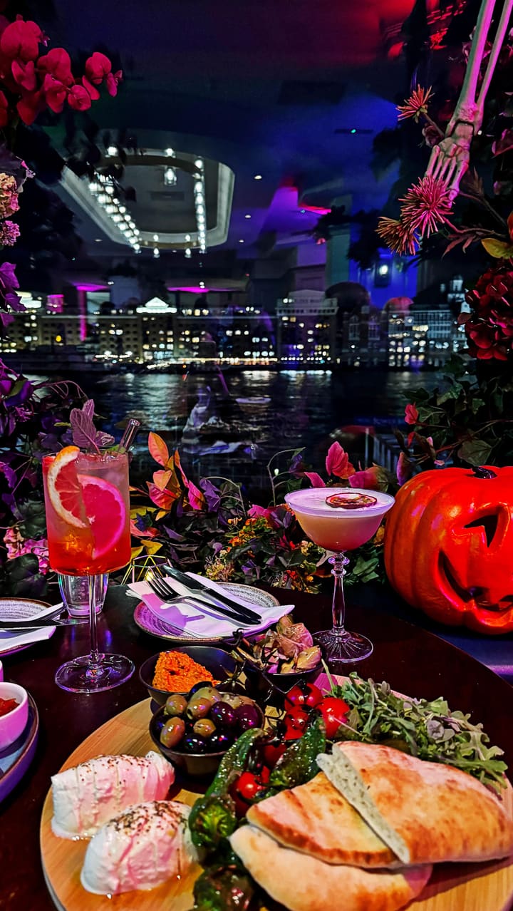 The video promotes a Halloween-themed event at the VU Hotel bar, located by Tower Bridge in London. It showcases the venue's spooky decorations, including pumpkins, skeletons, and floral arrangements, against the backdrop of the illuminated bridge at night. The video features various cocktails, such as a passionfruit martini, and food items including a burger and steak with fries. The atmosphere is presented as moody and stylish, with prominent red and purple lighting and glimpses of costumed staff and guests.