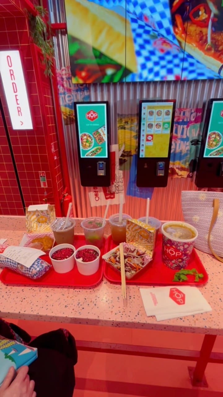 This video showcases Hop Vietnamese restaurant located at Bond Street station in London. The video starts with a touchscreen kiosk for placing orders, proceeds to show the overall ambiance of the restaurant including bright lights and hanging lanterns. Then, it shows the process of ordering and receiving the food, which includes chicken Pho, Viet slaw, and drinks like lemonade and bubble tea. Finally, it shows the food and drinks being enjoyed in the restaurant, with additional shots of the restaurant's interior design. The video has a vibrant and energetic feel throughout.