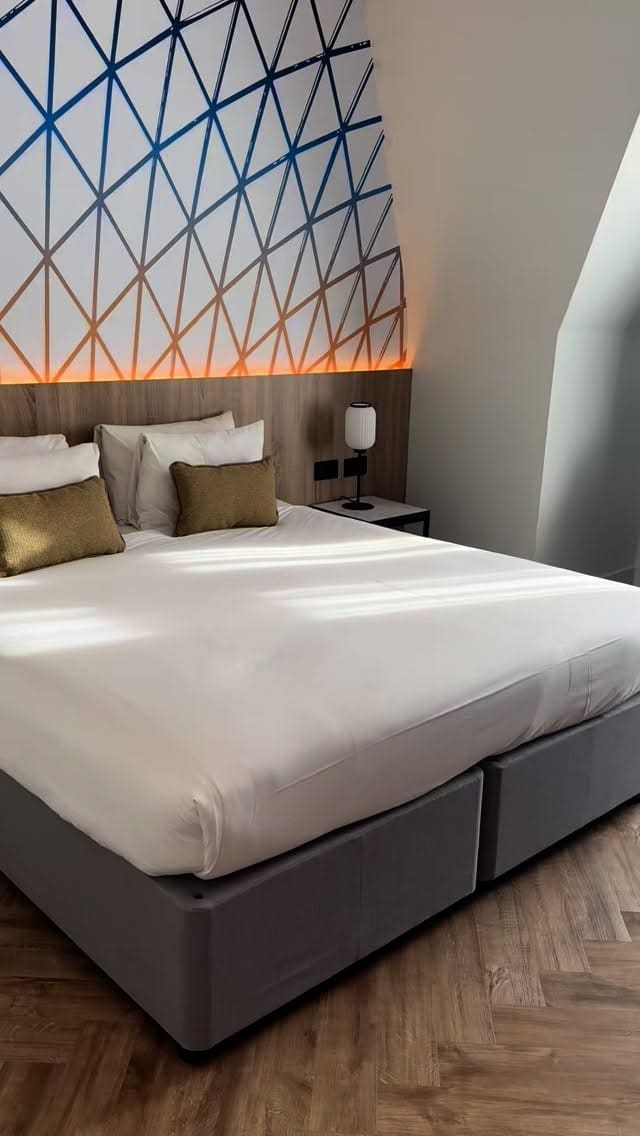 The video showcases a stay at the Thistle Hotel in Bloomsbury, London. It begins with shots of a modern, refurbished hotel room, including the bed and a stylish bathroom with a backlit mirror and a walk-in shower. Two women are shown enjoying the room in pajamas, drinking from mugs, and doing their skincare routine. One woman is seen opening the curtains to a bright window and later sitting by it with a drink. The video concludes with several shots of the hotel's extensive breakfast buffet, featuring hot items like bacon and sausages, pastries, cereals, fruits, and cheeses.