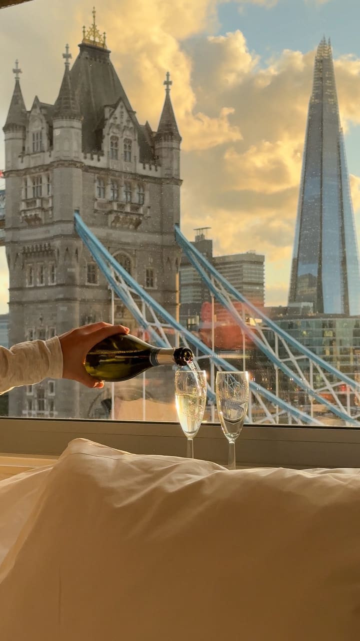 A montage of clips showing a luxurious hotel stay experience. The video features a person pouring champagne into two glasses on a windowsill with a stunning view of London's Tower Bridge and The Shard at sunset. It includes shots of two people toasting with their glasses and a person relaxing on a bed, enjoying the view with a glass of champagne. The overall mood is romantic and aspirational.
