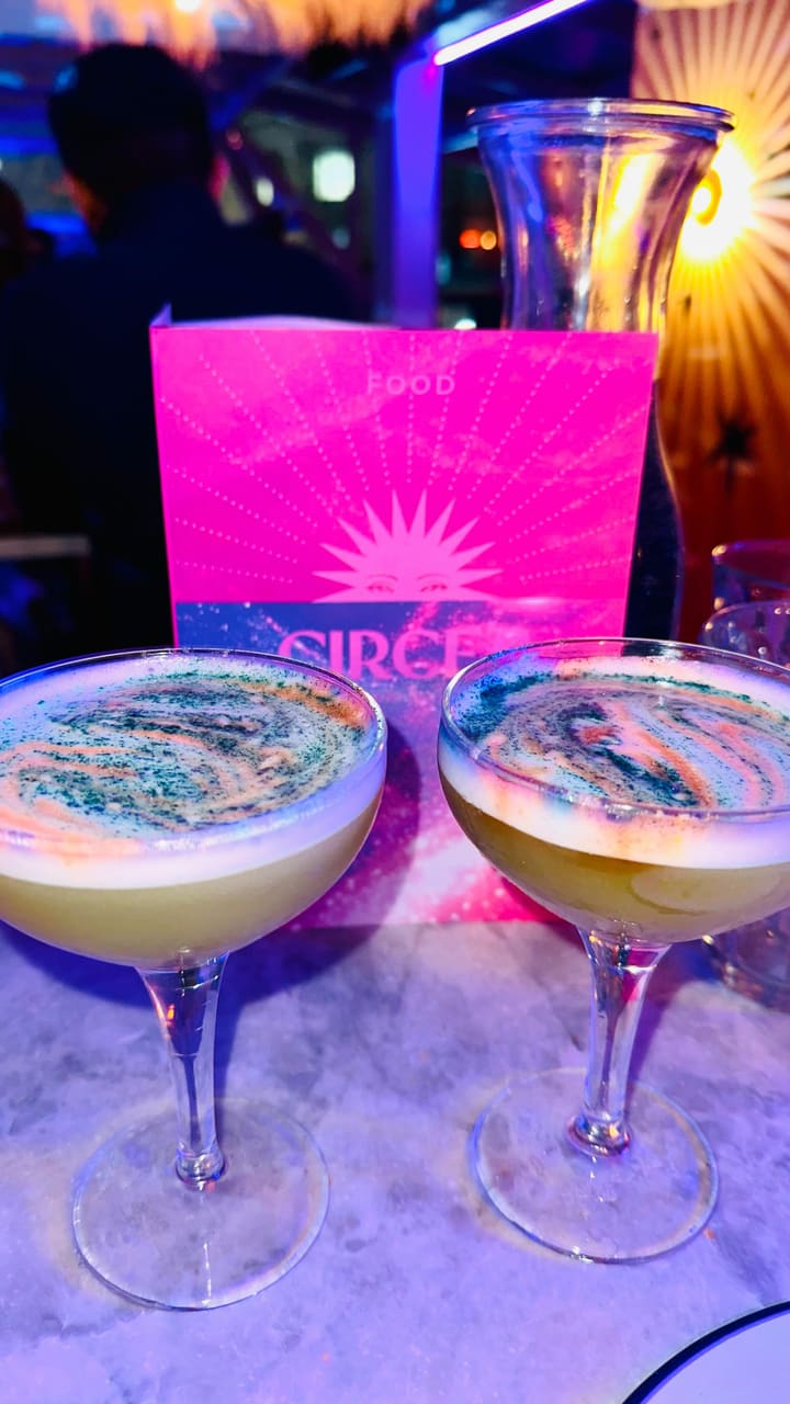 The video promotes Circe's Realm, a rooftop bar event in London. It begins with a shot of the entrance and a wall mural, followed by scenes of the bar's interior, featuring people socializing, a magician performing card tricks, and tarot card readings. The video showcases cocktails, sharing plates with grilled halloumi, salads, and mushroom dishes. It also shows a caricaturist drawing guests as mythical figures and a golden telephone prop. The main focus is on highlighting the unique experiences offered at the event, including cocktails, food, tarot readings, caricatures, and a chance to win prizes.