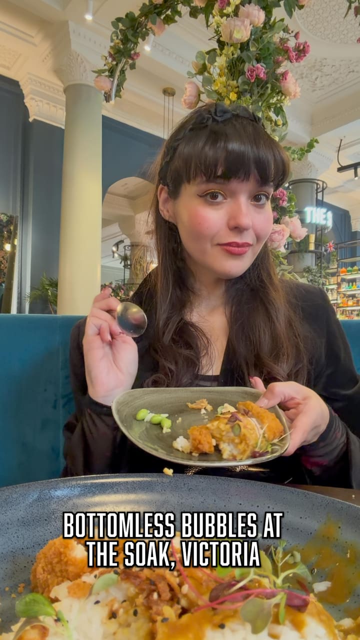 The video promotes a bottomless brunch at a restaurant called 'The Soak'. It features two women toasting with sparkling wine, followed by shots of various dishes including what appears to be calamari or spicy chicken bites, a chicken katsu curry, and pancakes topped with berries and cream. The video also shows sparkling wine being poured into glasses and the women enjoying their meal in the restaurant's aesthetically pleasing, flower-decorated interior. The final shot is of the restaurant's exterior sign, which is surrounded by a floral display.
