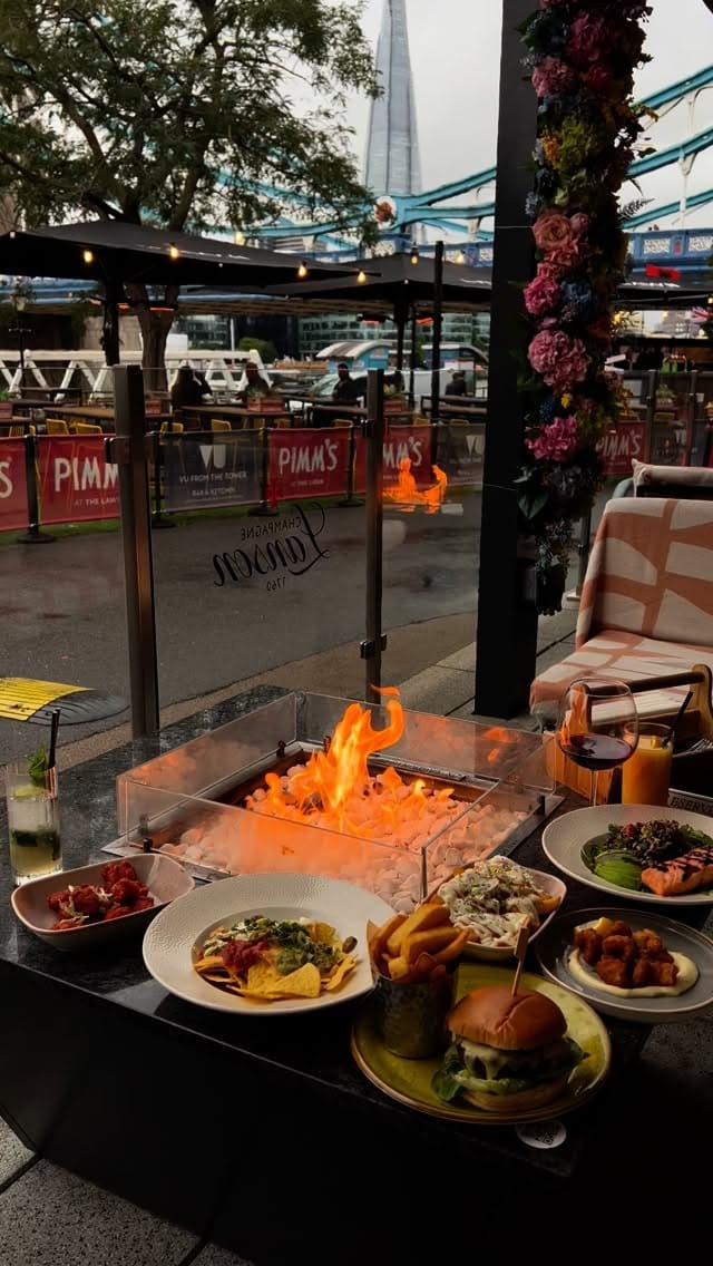 The video showcases an outdoor dining experience at a restaurant in London, with views of Tower Bridge and The Shard. A variety of dishes are displayed on a table with a central fire pit, including a cheeseburger with fries, nachos, popcorn chicken, truffled chips, and a salmon salad. The video features close-up shots of the food and people interacting with the dishes. Drinks shown include red wine, a mojito, and orange juice. The overall atmosphere is cozy and scenic.