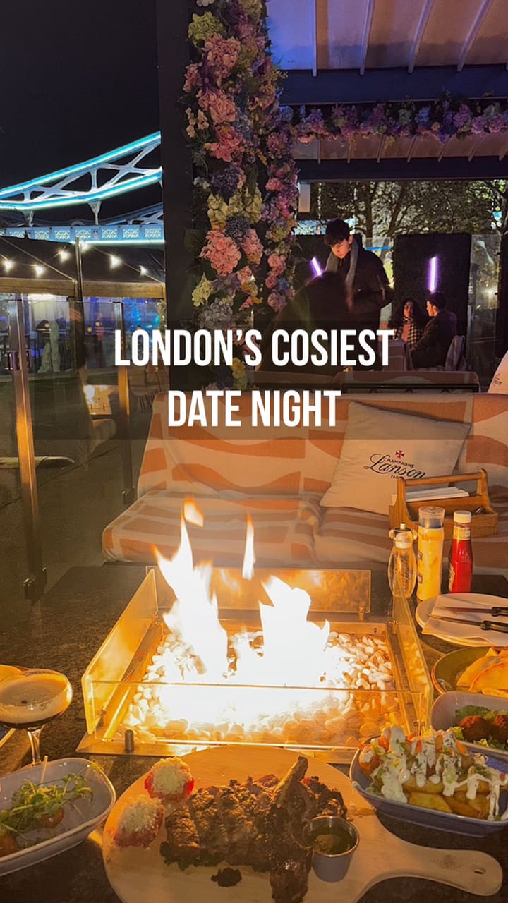 The video promotes a cozy date night experience at a restaurant named Vicinity, located by the River Thames with a clear view of Tower Bridge at night. The main feature is a private table with a fire pit. The video showcases various food and drink items, including two cocktails (an espresso martini and a tropical-style drink), a large tomahawk steak served sliced on a wooden board, loaded potato wedges, corn on the cob, meatballs, and pastries.