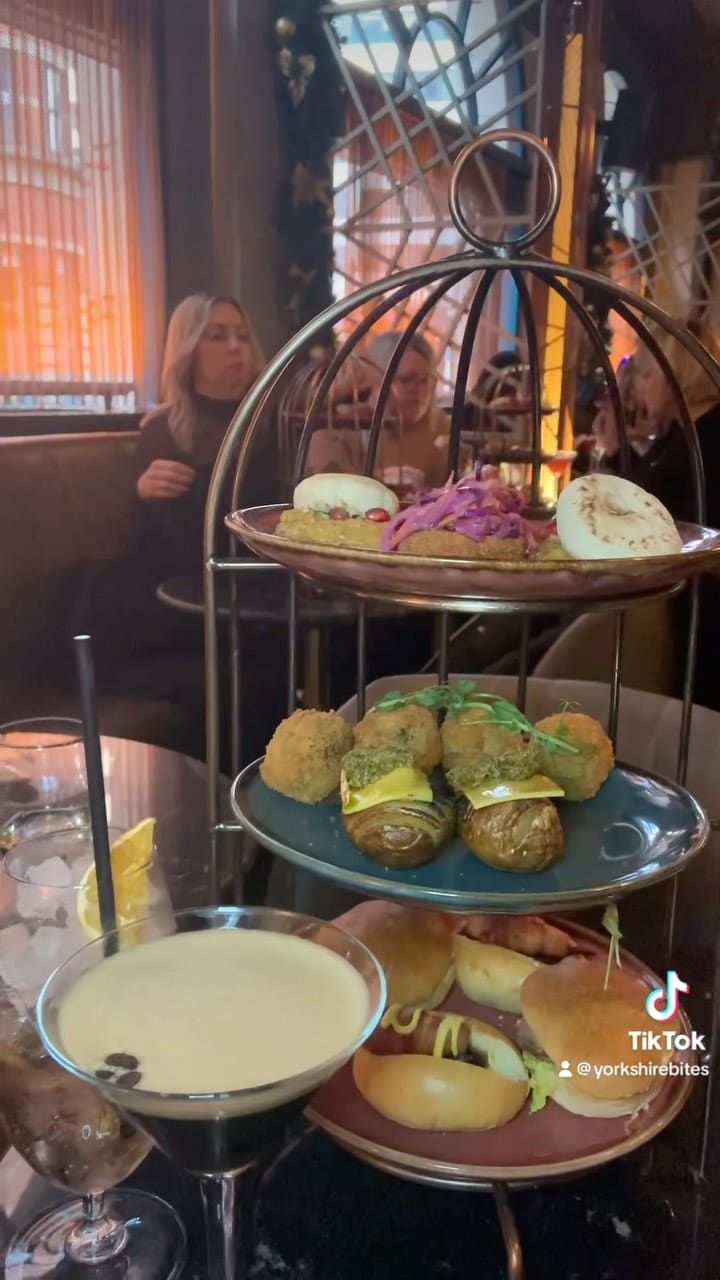 The video showcases a Christmas-themed bottomless brunch at Dirty Martini in Leeds. It features various cocktails (including a Christmas Martini and a White Christmas cocktail), and a selection of food items presented on a three-tiered stand.  These include mini pigs in blankets, turkey sliders, gouda and bacon donuts, flatbreads with hummus, roasted potato hasselback, and truffle mushroom arancini. The food items are presented as bite-sized portions, visually appealing and arranged on plates. No specific dietary information is provided. The primary focus is showcasing the food and drinks while interspersing snippets of a song.