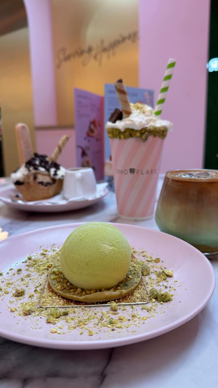 The video showcases Snowflake Gelato, a cafe offering various summer treats. It features a pistachio gelato dessert ('tennis ball' themed), coffee, and strawberry-themed drinks. The food presentation emphasizes pastel colors and visually appealing arrangements. No specific dietary information is provided. The main focus is presenting the cafe's summer menu items.