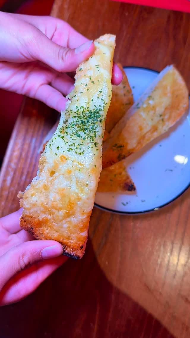 The video showcases a meal at a TGI Fridays restaurant. It begins with a close-up of a cheese pull from a piece of garlic bread. It then shows the exterior of the restaurant, followed by a wide shot of a table laden with various dishes. The dishes featured include fully loaded nachos, glazed salmon with mashed potatoes, corn on the cob, and onion rings, chicken tenders with fries, boneless wings, and two different chocolate desserts (a Mississippi Mud Pie and a chocolate fudge cake). The video also shows two cocktails and ends with shots of the restaurant's interior.