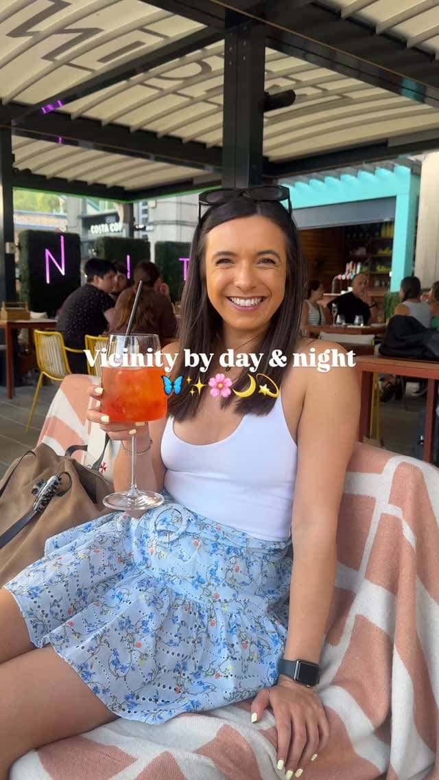 A video montage showcasing the Vicinity restaurant in London, transitioning from a daytime to a nighttime setting. The video features its outdoor terrace with prominent views of Tower Bridge. A variety of food and drinks are displayed, including Aperol Spritz-style cocktails, corn ribs, nachos, loaded potato wedges, a grilled steak with chips, a grilled chicken salad, a margarita, and an espresso martini. The atmosphere is highlighted, showing patrons enjoying their meals and the ambiance, which includes a fire pit in the evening.
